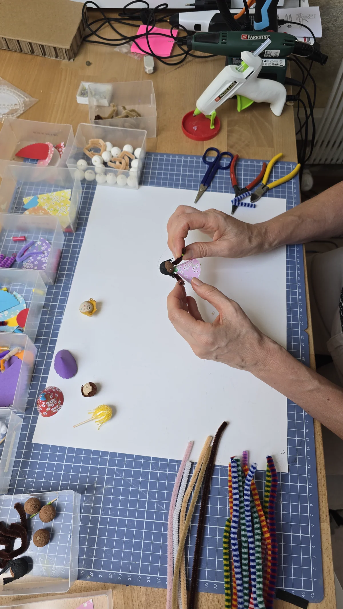 Mains de Clotilde assemblant un personnage du mobile : papiers de couleur, chenilles, boules de papier, outils d'atelier